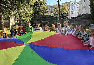 Dzieci z grupy trzeciej i czwartej stoją w ogrodzie przedszkolnym i podrzucają chustę animacyjną.