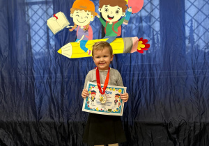 Dziewczynka stoi na tle dekoracji. Uśmiecha się, trzyma w rączkach dyplom. Na szyi ma założony medal.