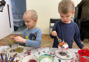 Dwóch chłopców ozdabia szklane bombki. Jeden z nich sypie brokat, drugi stempluje farbami.