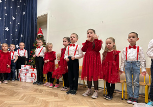 Dzieci z grupy trzeciej i czwartej stoją w półkolu. Śpiewają piosenkę ,,Skrzypi wóz".