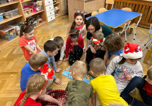 Dzieci siedzą w kole. Na środku koła siedzi mama chłopca. Kuba siedzi na kolanach mamy, która czyta książkę. Dzieci oglądają książkę.