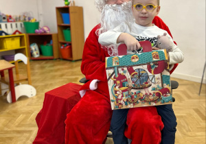 Chłopiec siedzi u Mikołaja na kolanie, Trzyma w ręku prezent.