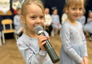 Dwie dziewczynki stoją na środku sali. Jedna z dziewczynek trzyma mikrofon i śpiewa piosenkę ,,Kocham Babcię".