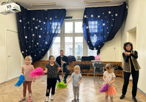 Przed Panem, który gra na saksofonie, stoją cztery dziewczynki i trzymają kolorowe pompony.