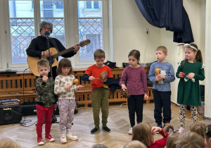 Dzieci stoją na środku sali i trzymają instrumenty, na których grają.