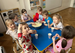 Dzieci siedzą przy stoliku i jedzą popcorn.