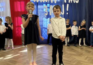 Dziewczynka i chłopiec stoją na środku. Dziewczynka trzyma mikrofon i mówi wiersz.