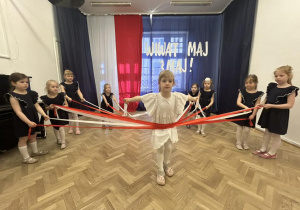 Dziewczynki trzymają wstążki, jedna z nich przechodzi przez wstążki.