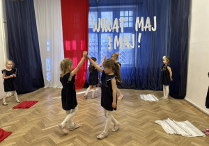 Dziewczynki stoją na środku sali, trzymają się za ręce.