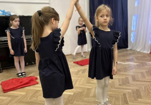 Dziewczynki stoją na środku sali, trzymają się za ręce.