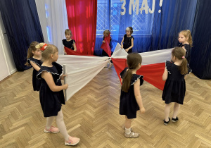 Dziewczynki trzymają dwie białe i czerwone chusty, tworzą na podłodze gwiazdę.