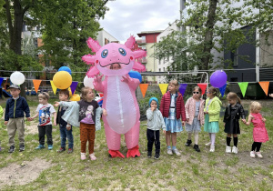 Dzieci ustawieni w kole tańczą z różowym dinozaurem.