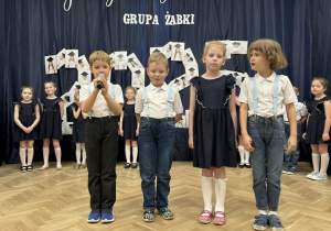 Czwórka dzieci stoi. Chłopiec trzyma mikrofon i mówi wiersz.