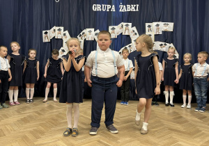 Trójka dzieci stoi na scenie. Dziewczynka trzyma mikrofon i mówi wiersz.
