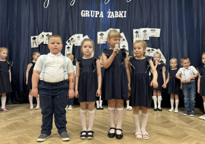 Czwórka dzieci stoi na scenie. Dziewczynka trzyma mikrofon i mówi wiersz.
