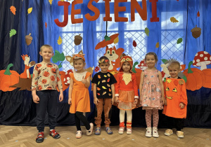 Dzieci z grupy III stoją w strojach jesiennych na tle dekoracji.