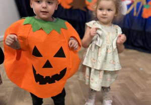 Dziewczynka i chłopiec stoją ubrani w strojach jesiennych, na tle dekoracji.