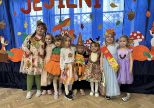 Dzieci z grupy IV stoją w strojach jesiennych na tle dekoracji.