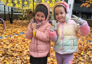 Dwie dziewczynki stoją w liściach. Jedna dziewczynka trzyma parasolkę.