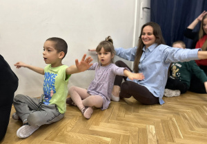 Dzieci wraz z rodzicami siedzą w kole i trzymają ręce na boku, jak samoloty.