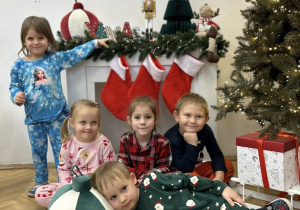 Dzieci z grupy II stoją, siedzą i leżą na tle dekoracji świątecznej.