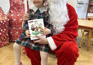 Dziecko siedzi na kolanie Mikołaja i odbiera prezent.