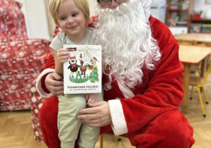 Dziecko siedzi na kolanie Mikołaja i odbiera prezent.