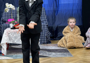 Chłopiec przebrany w garnitur i w białych pokazuje na podłogę.