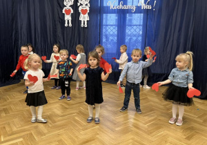 Dzieci stoją na tle dekoracji trzymają w rączkach czerwone serduszka.