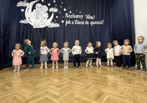 Dzieci stoją w półkolu na tle dekoracji.