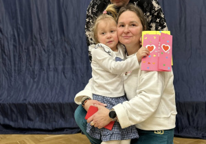 Babcia trzyma na kolanach wnuczkę i razem z dziadkiem uśmiechają się.