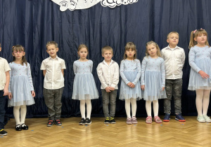 Dzieci z grupy IV stoją w półkolu na tle dekoracji.