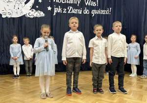 Trzech chłopców stoi na środku sali, dziewczynka mówi do mikrofonu.