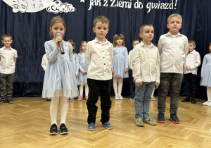 Trzech chłopców stoi na środku sali, dziewczynka mówi do mikrofonu.