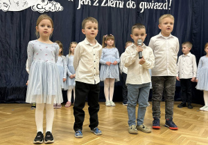 Trzech chłopców i dziewczynka stoją na środku sali, chłopiec mówi do mikrofonu.