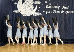 Dziewczynki stoją przy dekoracji, trzymając doniczki w górze.