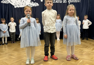 Dwie dziewczynki i chłopiec stoją na środku sali. Jedna z dziewczynek mówi wiersz.