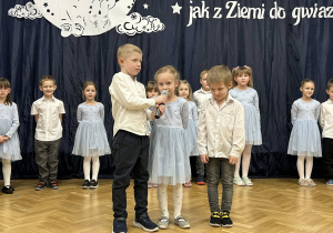 Dwóch chłopców stoi na środku sali, dziewczynka mówi do mikrofonu.