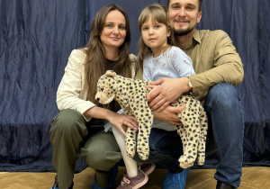 Dziewczynka siedzi na kolanach taty, obok kuca mama, wszyscy się uśmiechają.