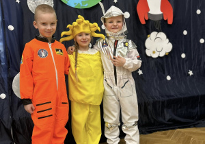 Dwóch chłopców i dziewczynka w kosmicznych strojach obejmują się i uśmiechają się.