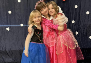 Trzy dziewczynki w strojach księżniczek przytulają się i uśmiechają się.