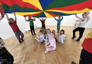 Część dzieci trzyma chustę animacyjną, reszta dzieci siedzi pod chustą.