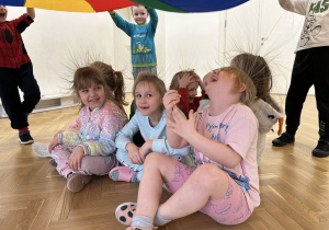 Dzieci siedzą pod chustą animacyjną i się uśmiechają.