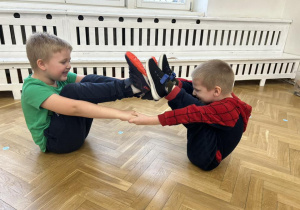 Chłopcy trzymają się za ręce, stykają się stopami.